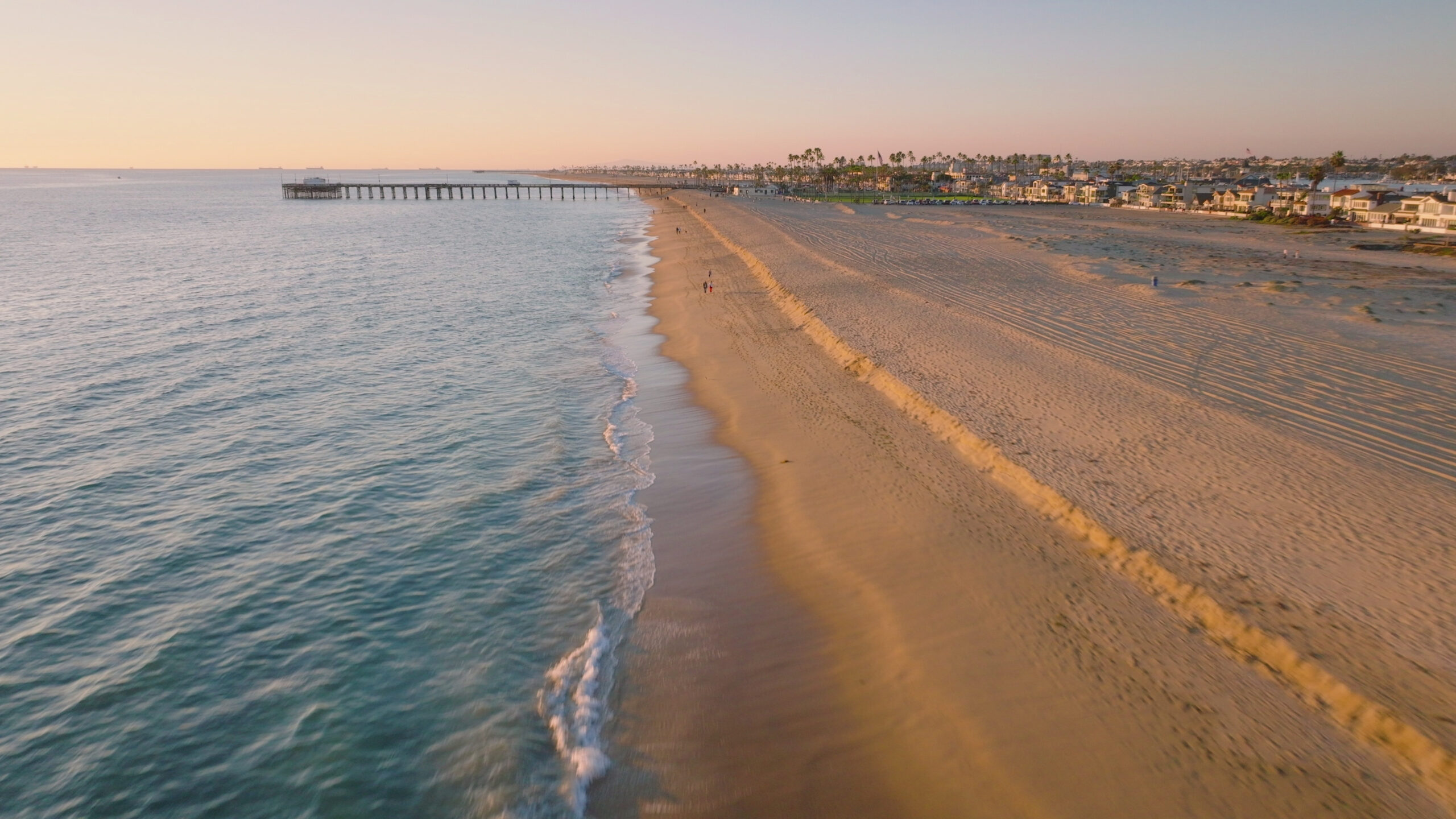 Aerial view of Newport Beach, California coastline at sunrise with pier and gentle waves.